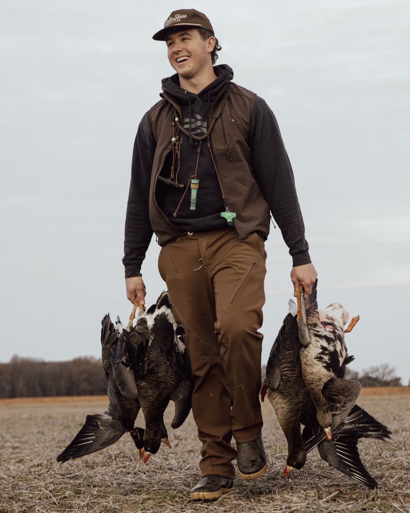 A smiling hunter walks out of the field carrying geese in both hands